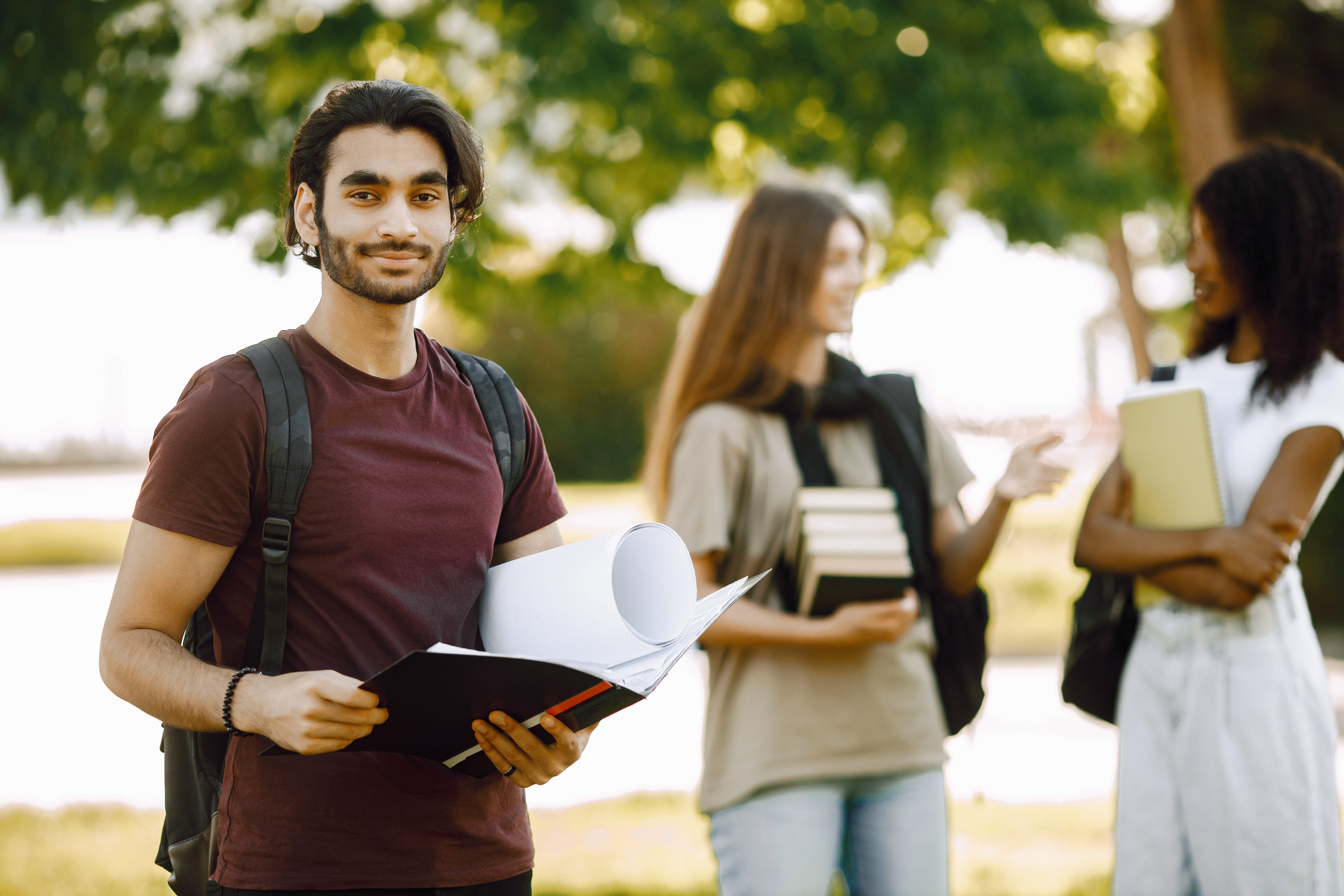 Focus on a indian boy who standing sepately. Group of international students standing together in park at university. Blurred african and caucasian girls talking outdoors.