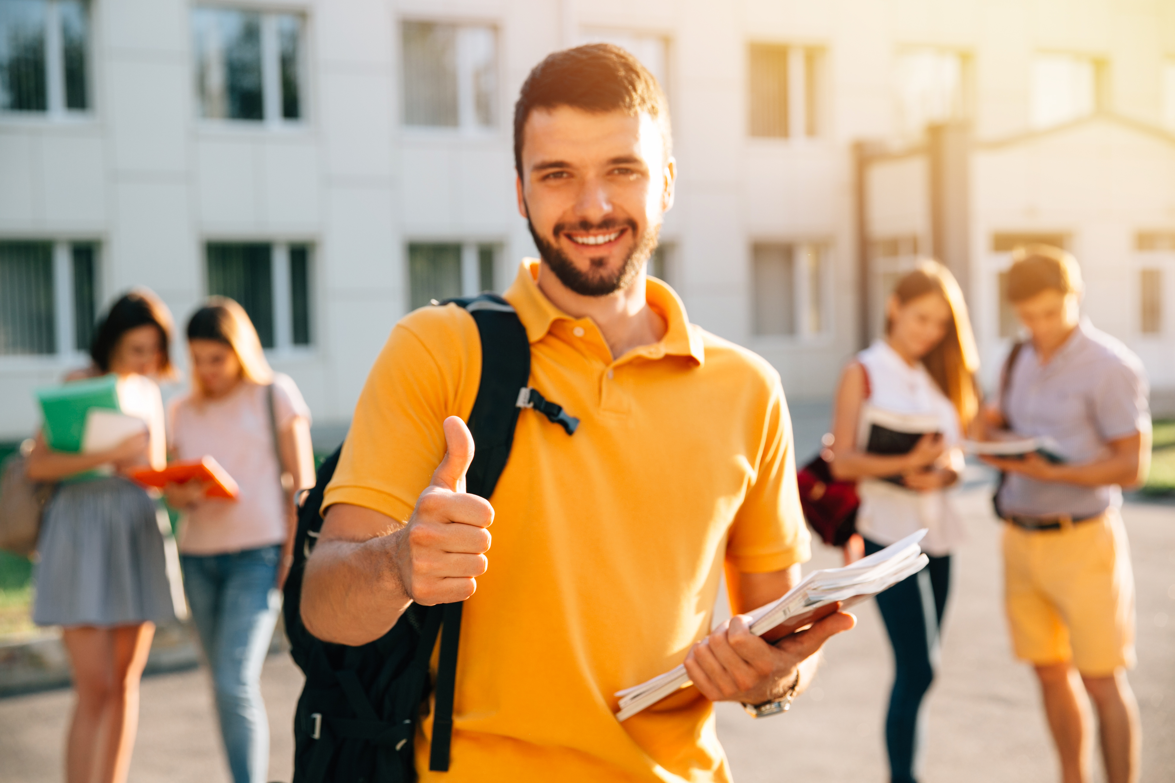 Young attractive smiling student showing thumb up outdoors on campus at the university. Selective focus